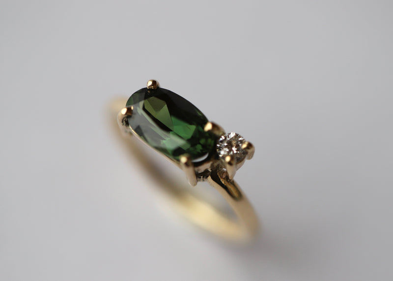 An elegant gold ring featuring an oval-cut green sapphire with a smaller diamond on the side, positioned on a plain white background.