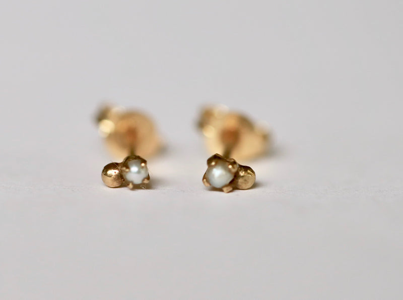 A pair of gold stud earrings featuring a small gold bead and a pearl set to the side, displayed on a white background.