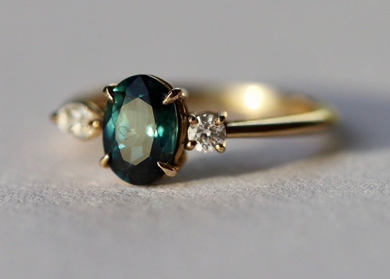 Gold ring with a teal sapphire and smaller diamonds on a light gray background