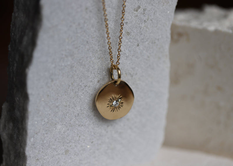 A gold necklace featuring a semi-round polished disc with a diamond set in a sunbeam setting, displayed against a stone surface.