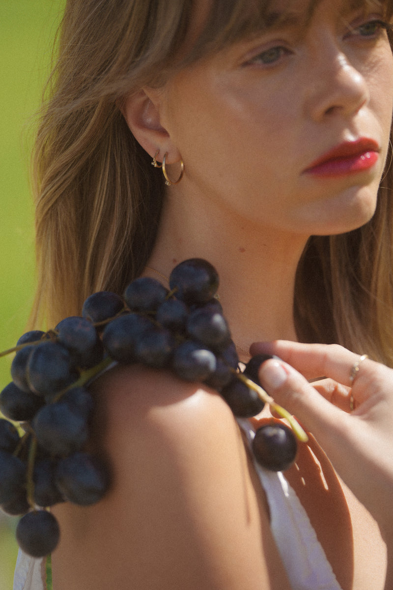 Model wears the Manahau hoops, gold organically shaped featuring a 2mm pearl claw set in the middle. Model looks away holding a bunch of grapes. 