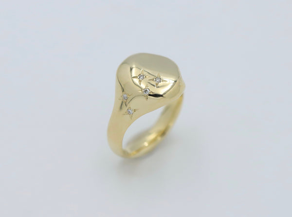 An oval-shaped yellow gold signet ring with a row of scattered diamonds set on one side, displayed against a white background.