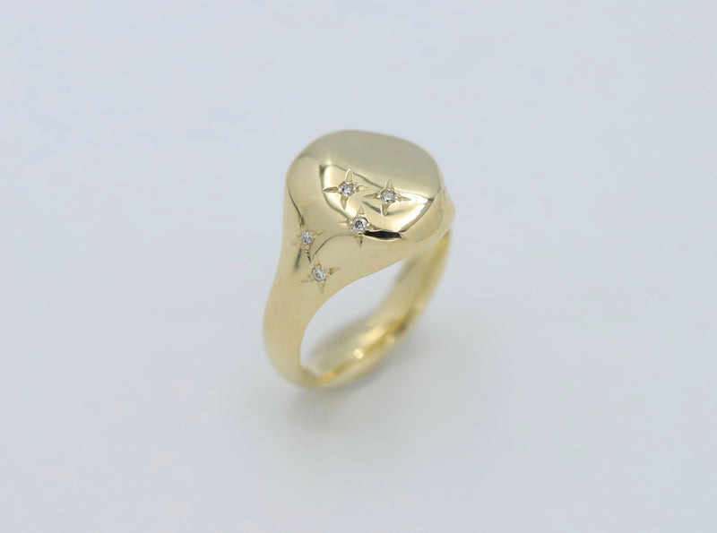 An oval-shaped yellow gold signet ring with a row of scattered diamonds set on one side, displayed against a white background.